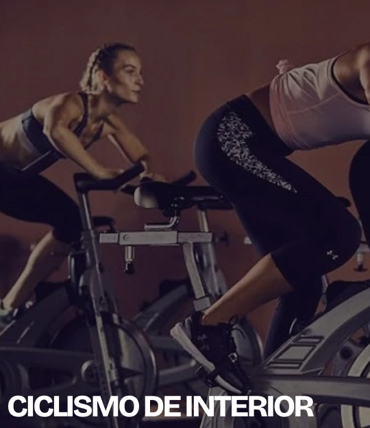 Dos mujeres pedalean en bicicletas estáticas durante una clase de ciclismo de interior, con el texto “CICLISMO DE INTERIOR” en la parte inferior.