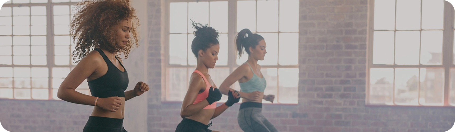 Tres mujeres con ropa deportiva trotan en fila dentro de un gimnasio luminoso, con grandes ventanas y una pared de ladrillo al fondo.